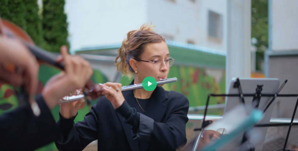 Eine junge Musikerin spielt konzentriert Querflöte, während im Vordergrund verschwommen eine Geige zu sehen ist. In der Mitte des Bildes befindet sich ein Play-Button, der auf ein Video hinweist.